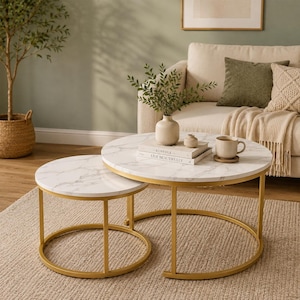 May include: A set of two round nesting coffee tables featuring white faux marble tops with grey veining and gold-colored metal frames. The larger table holds a white ceramic vase with greenery, a small white bowl, and a book titled KINFOLK.