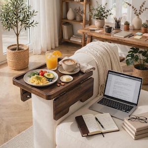 Puede incluir: Una bandeja de madera oscura para el brazo del sofá con superficie rectangular y laterales abatibles. Sostiene un plato blanco con huevos revueltos, aguacate y tomates, un vaso de zumo de naranja, una taza de café y mantequilla.