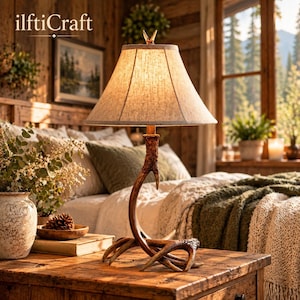 May include: A table lamp featuring a base crafted from brown deer antlers and a beige fabric lampshade. The lamp emits a warm glow, resting on a wooden surface. The text "ilftiCraft" is visible in the upper left corner of the image.