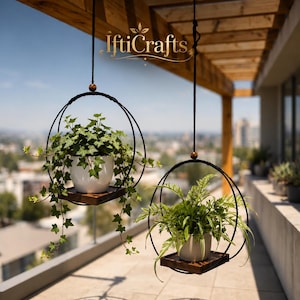 Puede incluir: Dos soportes colgantes para plantas con marcos circulares de metal negro y bases cuadradas de madera oscura. Cada soporte contiene una maceta de cerámica blanca con follaje verde, suspendida por cordones negros con cuentas de madera. El texto IftiCrafts es visible.