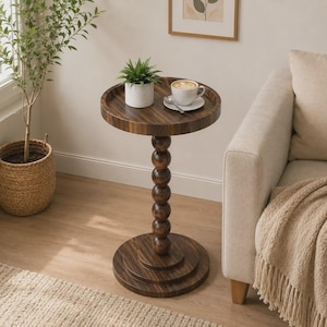 May include: A round dark wood side table with a raised edge and a central pedestal base composed of stacked wooden spheres. The table features a smooth, polished finish and rests on a circular base. A small potted plant and a white coffee cup sit on top.
