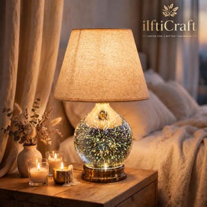 May include: A table lamp with a beige fabric conical shade and a spherical glass base featuring a multi-colored 3D firework light effect. The lamp sits on a metallic base, emitting a warm glow that illuminates the surrounding space.