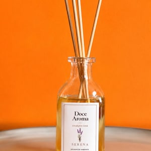 Pode incluir: Um frasco difusor de vidro transparente com líquido dourado e quatro varetas de madeira. O rótulo branco apresenta o texto Doce Aroma, Coleção Casa, Serena, Difusor de Ambiente, 100ml e uma pequena ilustração de lavanda.