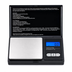 May include: A black digital pocket scale with a silver weighing platform and a blue backlit LCD screen. The display shows 0.0, with text indicating a 500g capacity and 0.1g graduation. The lid features printed operating instructions in English.