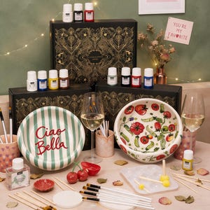 Könnte beinhalten: Ein Bastelset mit zwei schwarzen Boxen mit goldenen Blumenmustern, Farbdosen, Pinseln, einer weißen Palette und zwei Keramikschalen. Eine Schale ist grün gestreift mit dem Text Ciao Bella, die andere zeigt ein bemaltes Tomaten- und Erbsenmotiv.