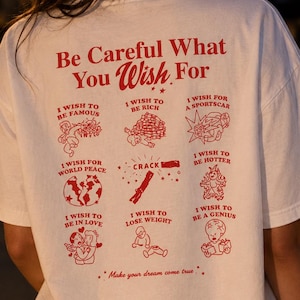 Könnte beinhalten: Ein weißes kurzärmeliges T-Shirt mit rotem Grafiktext und Illustrationen auf der Rückseite. Der Text lautet Be Careful What You Wish For, gefolgt von neun Illustrationen mit Bildunterschriften wie I Wish To Be Famous, I Wish To Be Rich, I Wish For A Sportscar, I Wish For World Peace, Crack, I Wish To Be Hotter, I Wish To Be In Love, I Wish To Lose Weight, I Wish To Be A Genius und Make your dream come true.