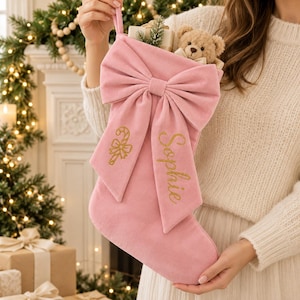 May include: A soft pink velvet Christmas stocking featuring a large decorative bow on the front. The stocking is embroidered with a gold glitter candy cane and the name Sophie in gold script. A small plush teddy bear sits inside the top.