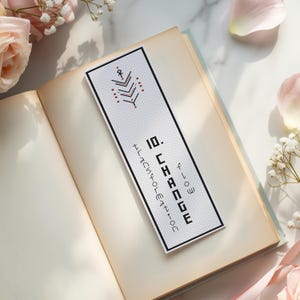 May include: A rectangular white bookmark featuring black cross-stitch embroidery. The design includes a geometric arrow symbol at the top and the text 10. CHANGE, flow, and TRANSFORMATION printed in a vertical orientation with a black border.