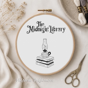 May include: A circular wooden embroidery hoop containing white fabric with a black cross-stitch design. The design features the text The Midnight Library above a stack of two books topped with a lit oil lamp, all rendered in black thread.