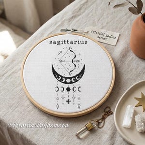 May include: A circular wooden embroidery hoop containing white fabric with a black cross-stitch design of a Sagittarius bow and arrow, a crescent moon, and hanging moon phases and stars. A paper tag reads celestial zodiac series.