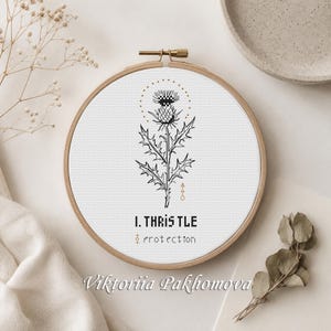 Pode incluir: Um bastidor de bordado circular de madeira contendo tecido branco com um design em ponto cruz preto de uma planta de cardo. Acima do cardo há um círculo pontilhado e abaixo o texto I. THRISTLE e a palavra protection em preto.