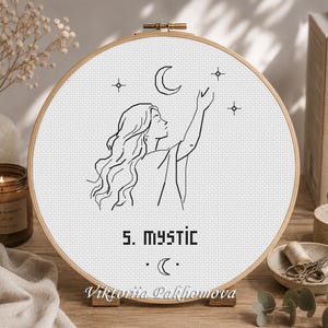 Peut inclure: Un tambour à broder circulaire en bois contenant une toile Aida blanche avec un motif au point de croix noir représentant une personne tendant la main vers un croissant de lune et des étoiles. Le texte 5. MYSTIC est brodé sous la figure.