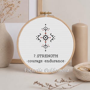 May include: A circular wooden embroidery hoop containing white fabric with a cross-stitch design. The pattern features a black and red geometric symbol above the text 7. STRENGTH and courage endurance. The name Viktoriia Pakliomova is printed below.
