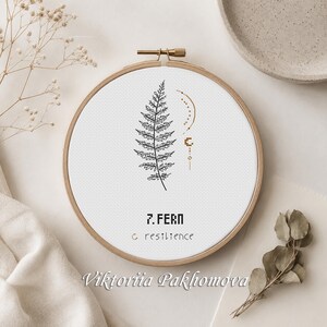 May include: A circular wooden embroidery hoop containing white fabric with a black cross-stitch design of a fern leaf, a small gold crescent moon, and the text 7. FERN and resilience in black and gold lettering centered on the fabric.