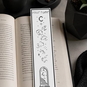 May include: A rectangular white bookmark featuring black cross-stitch embroidery. The design includes the text 'your name' at the top, followed by celestial motifs like stars, clouds, and a crescent moon, ending with an arched doorway graphic.