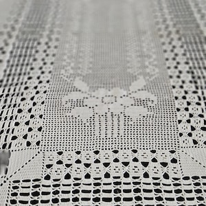 Könnte beinhalten: Ein rechteckiger weißer Häkel-Tischläufer mit einem komplizierten Durchbruchmuster, einem zentralen Blumenmotiv und einem dekorativen Bogenrand. Die feine Handarbeit zeigt ein sich wiederholendes geometrisches Design.