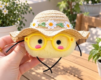 May include: A round, yellow plush toy featuring black embroidered eyes and pink cheeks, wearing bright yellow plastic glasses and a woven straw hat decorated with a band of white daisy flowers with green leaves and a thin black chin strap.