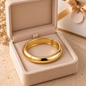 Può includere: Un bracciale rigido incernierato color oro lucido con una superficie liscia e arrotondata. Il bracciale è esposto all'interno di un portagioie aperto foderato in velluto beige, con un meccanismo di chiusura visibile sul lato dell'anello.