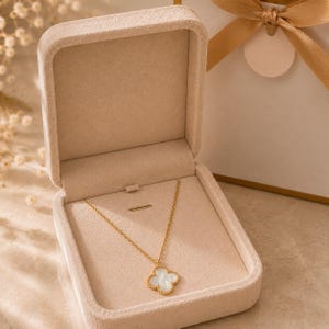 May include: A gold-toned necklace featuring a four-leaf clover pendant with a white iridescent inlay, presented inside an open, beige velvet-lined jewelry box. The necklace has a delicate chain and is displayed against a neutral-colored interior.