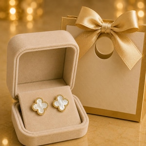 Puede incluir: Un par de pendientes de botón dorados con forma de trébol de cuatro hojas, incrustaciones de nácar blanco y bordes perlados, presentados en un joyero de terciopelo beige abierto junto a una bolsa de regalo a juego con un lazo de satén dorado.