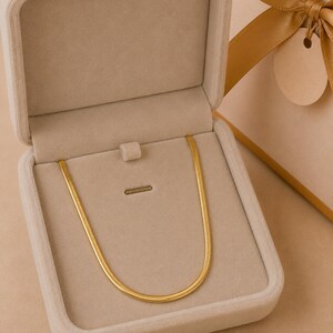 Puede incluir: Un collar de cadena de espiga en tono dorado que descansa dentro de un joyero forrado de terciopelo beige. El collar presenta un diseño de eslabones planos y flexibles con un acabado pulido. El joyero está abierto, mostrando el collar.