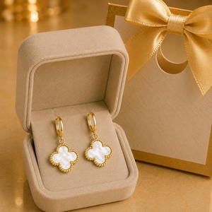 Puede incluir: Un par de pendientes de aro dorados con colgantes de trébol de cuatro hojas en color blanco y bordes perlados. Los pendientes se presentan dentro de un joyero beige forrado de terciopelo, junto a una caja de regalo con un lazo de satén dorado.