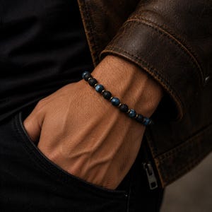 Puede incluir: Una pulsera de cuentas que alterna cuentas redondas de piedra de lava negra porosa y cuentas de piedra azul veteada lisa, acentuadas con pequeños espaciadores metálicos. La pulsera se lleva en la muñeca sobre una camisa oscura.
