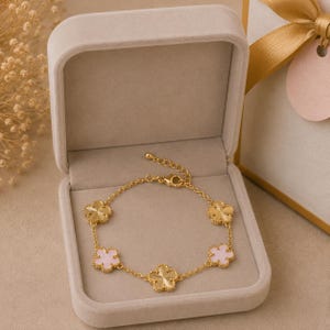Può includere: Un bracciale a catena color oro con cinque ciondoli alternati a forma di fiore, di cui due con finitura dorata testurizzata e tre con un inserto in smalto rosa chiaro liscio, chiuso da un fermaglio a moschettone.
