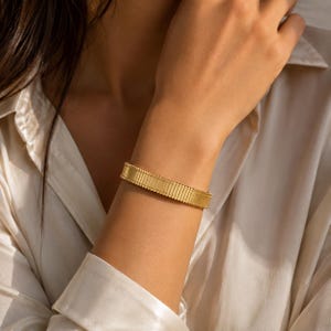 Peut inclure: Un bracelet manchette doré présentant une texture côtelée verticale répétitive sur toute la bande. Le bracelet a une largeur constante et une finition polie, avec de petits détails perlés le long des bords supérieur et inférieur de la bande.