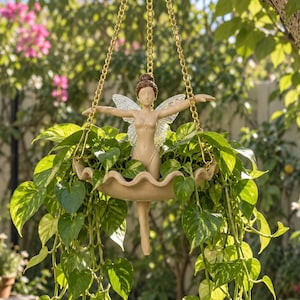 May include: A hanging planter featuring a beige fairy figure with outstretched arms and translucent wings, positioned in the centre of a scalloped bowl. The planter is suspended by gold-toned metal chains and contains a lush green pothos plant.