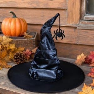 May include: A black witch hat featuring a wide brim and a pointed, slightly curved crown. A small, black, glittery spider ornament hangs from the tip of the hat by a thin black string, dangling against the side of the fabric cone.