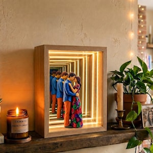 Puede incluir: Una caja de espejo infinito de madera rectangular con una tira de luz LED interna y una figura de una pareja abrazada. La persona de la izquierda viste camisa azul y pantalones naranjas, mientras que la de la derecha lleva un vestido floral.