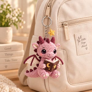 May include: A crochet dragon keyring in light pink with dark pink accents, featuring large purple eyes, small wings, and a brown book with a yellow moon and star design. A small yellow crochet star charm hangs from the silver metal keyring.