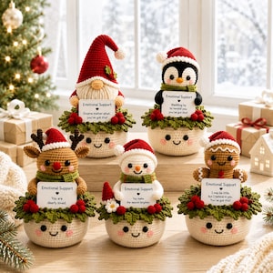 May include: Five crocheted figures in pots, including a gnome, penguin, reindeer, snowman, and gingerbread person. Each holds a white sign with the text Emotional Support and a unique message, surrounded by green leaves and red berries.