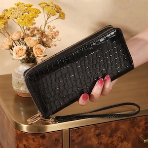 May include: A black rectangular wallet with a glossy crocodile-embossed texture, featuring a gold-tone metal zipper closure along the top and side, and a matching black wrist strap attached to the side of the wallet.