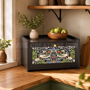 May include: A matte black bread box featuring a front panel with a colorful botanical pattern of birds, strawberries, and foliage. The box has a single gold-toned circular knob and sits on a wooden countertop next to a small vase with green leaves.