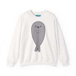 Può includere: Una felpa bianca a girocollo con una grande grafica centrale raffigurante una foca grigia che tiene in mano un piccolo pesce blu. La foca ha un design semplice e arrotondato con occhi, baffi e coda neri. Un'etichetta blu è visibile sul colletto.