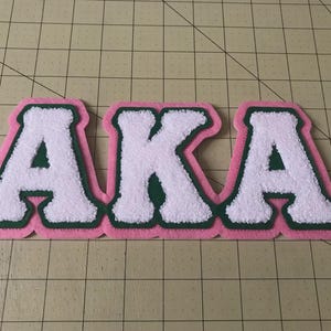 May include: A chenille patch featuring the letters AKA in white, outlined in dark green, and bordered by a light pink felt backing. The letters have a textured, fuzzy surface, and a small registered trademark symbol is visible on the final letter.