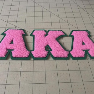 May include: A chenille patch featuring the letters AKA in bright pink with a dark green felt border. The letters have a textured, fuzzy surface and are connected in a single piece, designed for application onto fabric surfaces as a decorative accent.