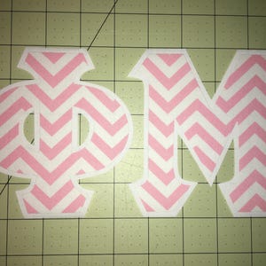 May include: Two fabric appliques: a Greek letter Phi and the letter M. Both are white with a pink chevron pattern. The Phi is a stylized, rounded shape. The M is a capital letter with a pointed top.