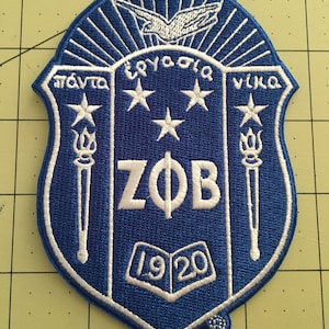 May include: A blue and white embroidered patch with the letters "ZOB" in white, surrounded by four white stars and the text "πάντα ἐργάζομαι νίκα" in white. The patch also features a white bird with outstretched wings and the year "1920" in white.