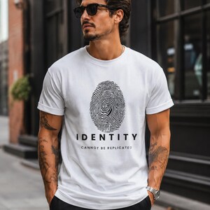 May include: A white short-sleeved t-shirt featuring a black graphic of a fingerprint centred on the chest. Below the fingerprint, the word IDENTITY is printed in bold black capital letters, followed by the text CANNOT BE REPLICATED.