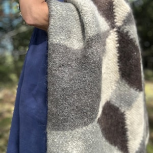 Könnte beinhalten: Eine dicke, strukturierte Wolldecke mit einem geometrischen Rautenmuster in den Farben Anthrazit, Hellgrau und Creme. Der Stoff hat eine flauschige, gefilzte Optik mit sichtbaren Fasern und einem Fransenrand an der unteren Kante.