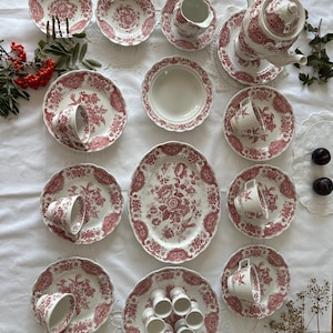 Pode incluir: Um conjunto de chá em cerâmica branca com um padrão floral vermelho. O conjunto inclui um bule, uma leiteira, um prato oval, vários pratos, pires, chávenas e uma coleção de pequenos suportes para ovos, decorados com motivos botânicos.