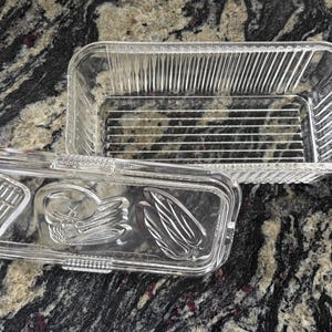 May include: A clear glass rectangular butter dish with a matching lid. The base features vertical ribbed sides and a horizontal ribbed bottom. The lid is embossed with three distinct vegetable designs, including a stalk of asparagus and corn.