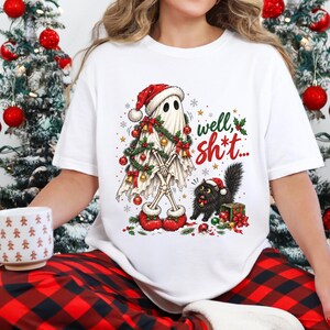 May include: A white t-shirt featuring a graphic of a ghost wearing a Santa hat and wrapped in Christmas garland, standing next to a black cat with a Santa hat and a gift box. The text "well, sh*t..." is printed in red and green script.