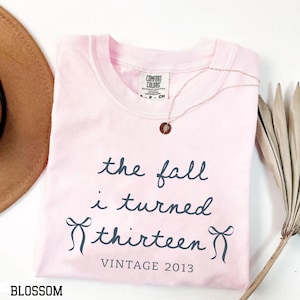 Puede incluir: Una camiseta de manga corta de color rosa claro con texto gris oscuro que dice "the fall i turned thirteen" y "VINTAGE 2013" junto a dos ilustraciones de lazos. La prenda incluye una etiqueta Comfort Colors y un collar dorado.