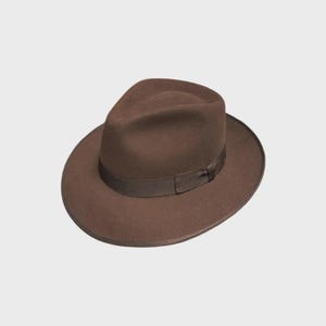 May include: A dark brown felt fedora hat featuring a pinched crown and a wide, structured brim. The hat is accented with a matching dark brown grosgrain ribbon band that encircles the base of the crown, providing a clean and uniform appearance.