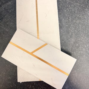 May include: Two rectangular white tiles with a subtle marbled texture and a single thin gold metallic line running across the surface. The tiles are arranged in an overlapping pattern, showcasing the clean geometric design and the contrast between the white marble-like finish and the reflective gold inlay.