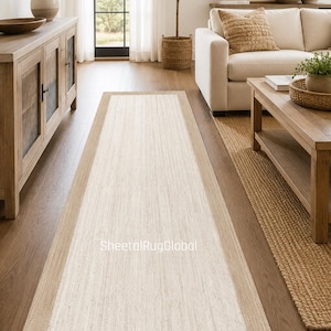 Può includere: Un tappeto passatoia rettangolare con una sezione centrale tessuta color bianco sporco, delimitata da una cornice color marrone chiaro più ampia. Il tappeto ha un aspetto testurizzato in fibra naturale con la scritta SheetalRugGlobal in bianco.